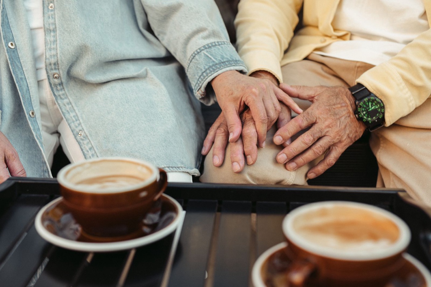 Hands of a couple in their fifties resting on each other at a café terrace, two cappuccinos in the foreground — dating after 50 in Luxembourg